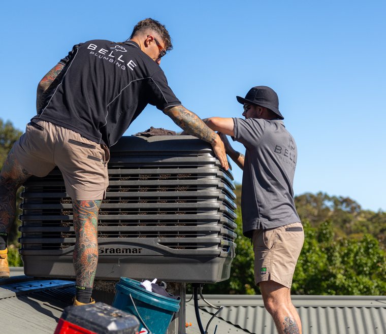 Two technicians lifting a replacement evaporative cooler into place